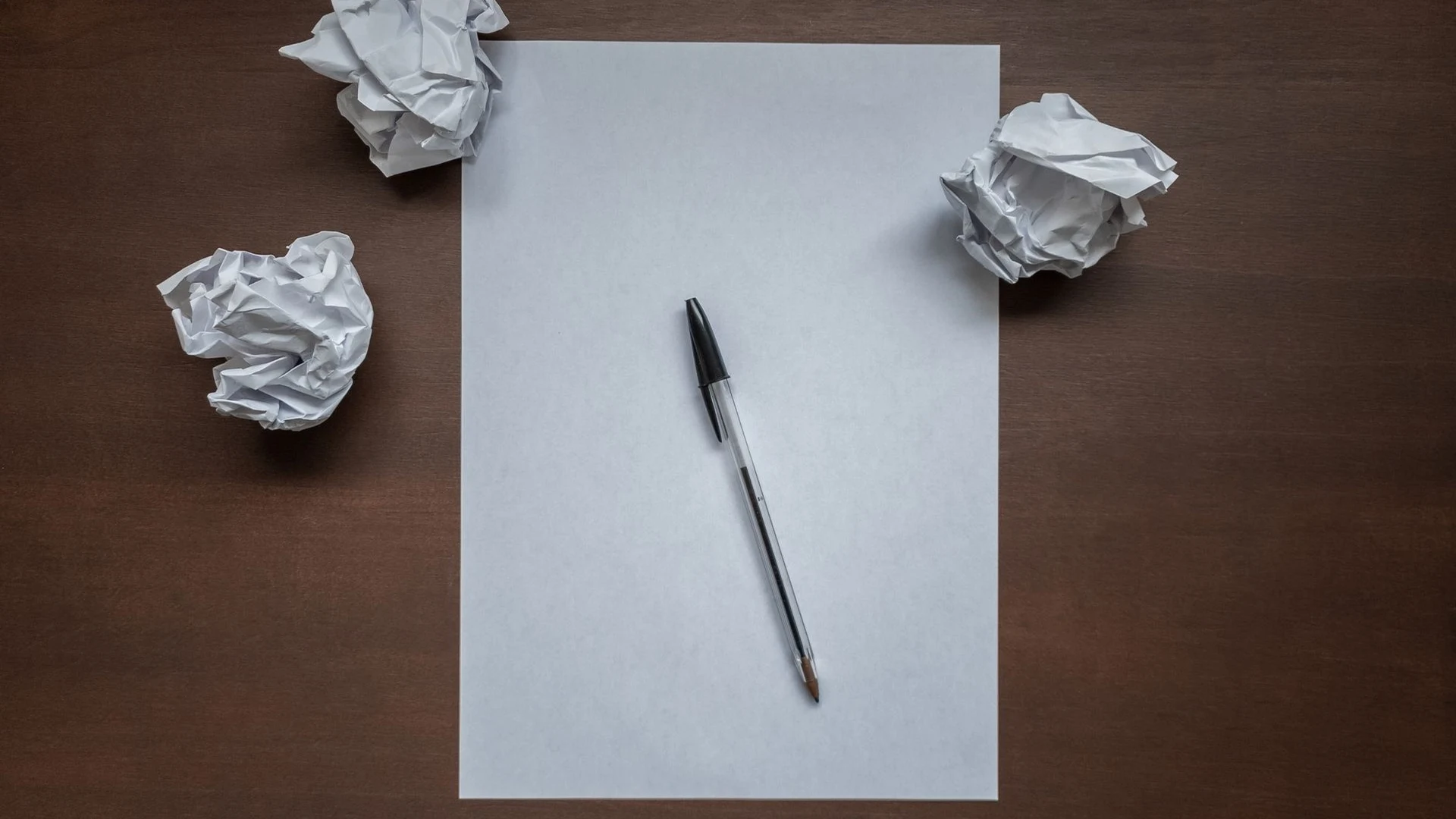 Blank sheet of paper and a pen on a wooden desk surrounded by crumpled paper balls