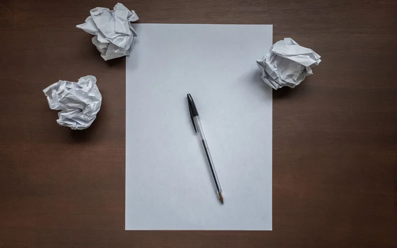 Blank sheet of paper and a pen on a wooden desk surrounded by crumpled paper balls
